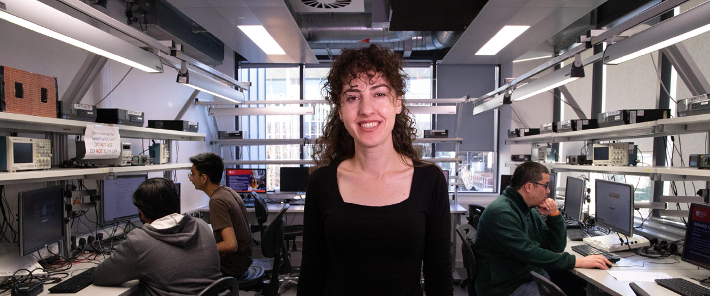 A young woman stands in a computer maintenance workshop looking directly into the camera