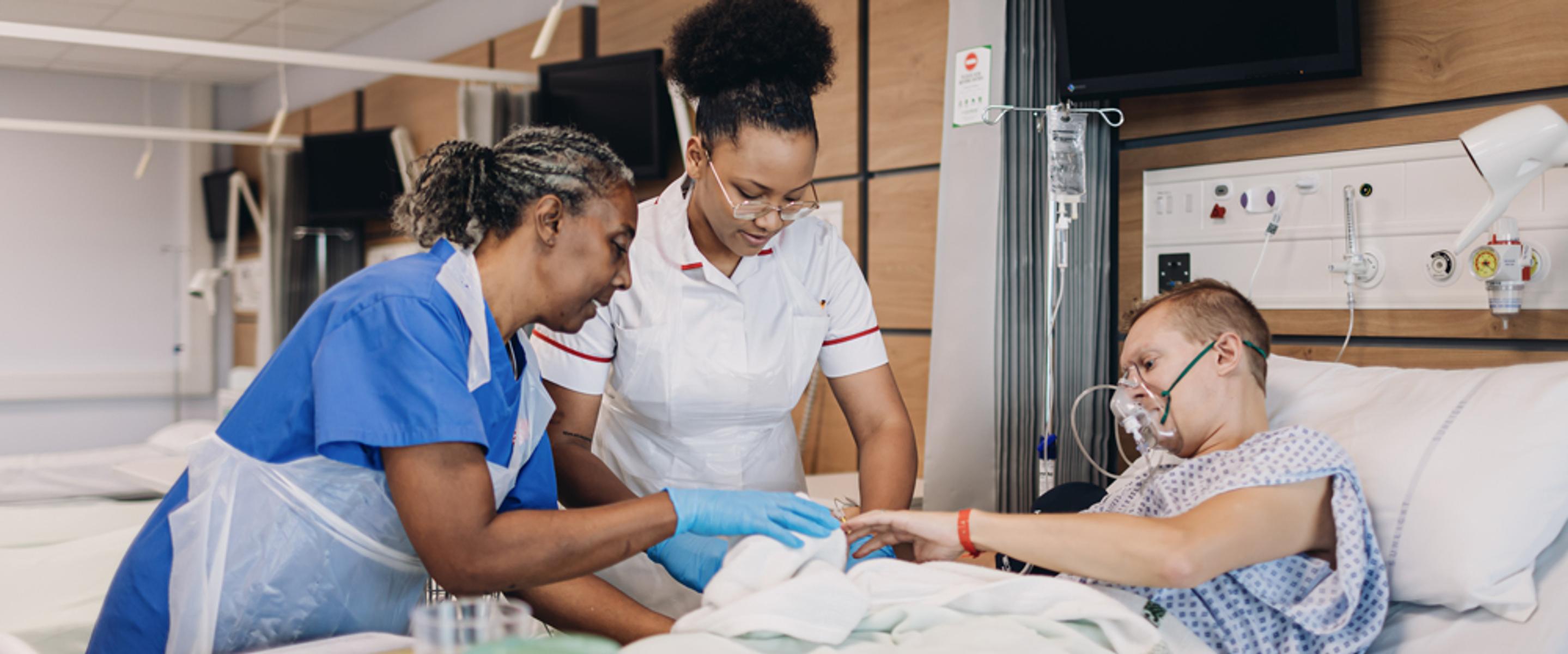 Two nurses undertaking a procedure on a patient wearing a mask and lying in bed