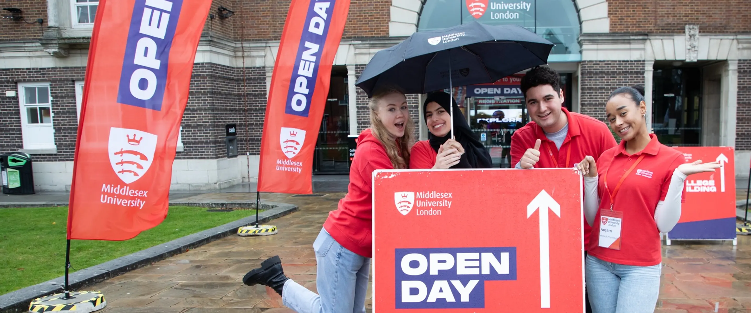 Four smiling students holding an umbrella, behind a sign pointing forwards to open day registration