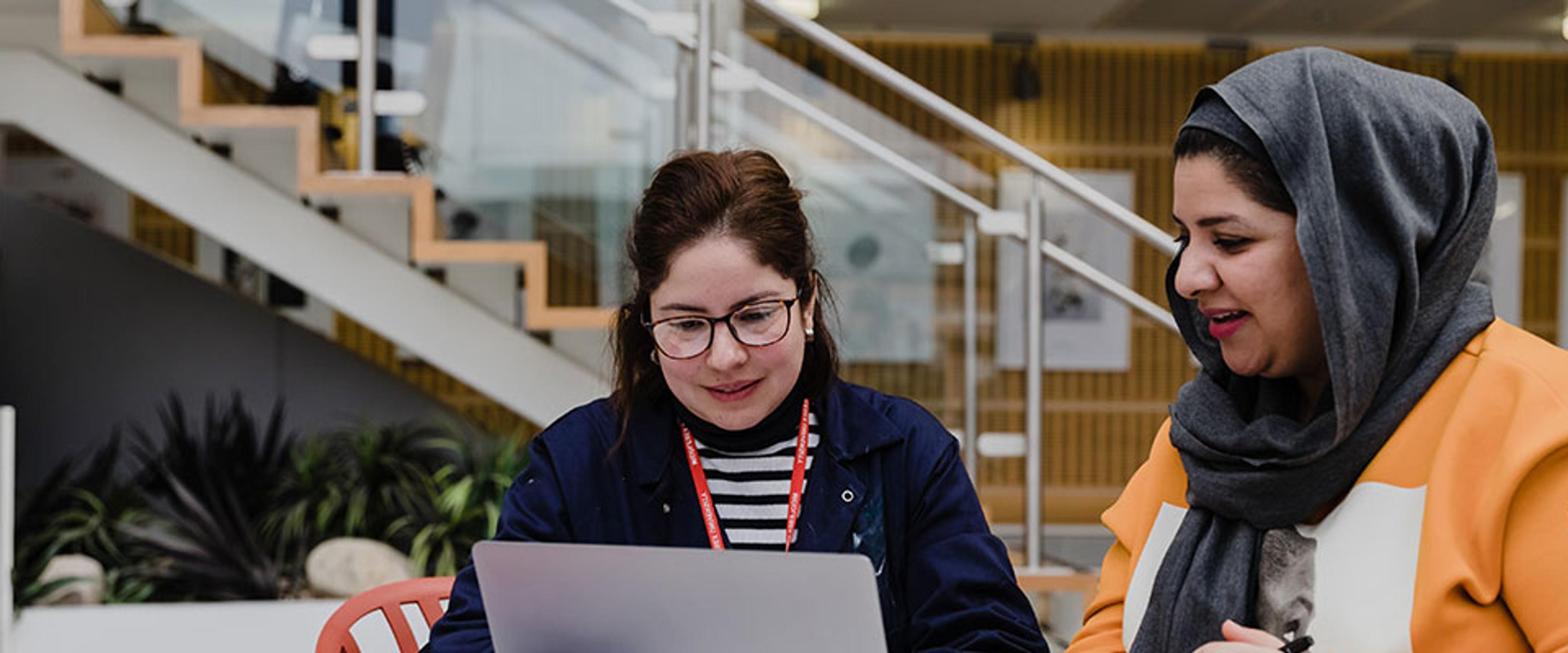 Two women sitting at a table with a laptop, discussing work projects and sharing ideas.