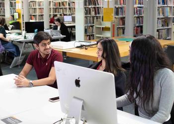 A group of students sit chatting in a work area with a desktop computer