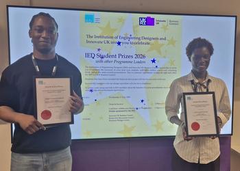 Two award winners holding certificates, with a screen behind them displaying their prize.