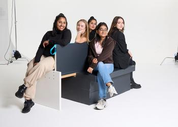 A group of five women sitting on a bench against a white photographic studio backdrop