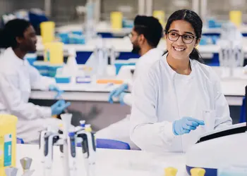 A smiling woman works in a science lab with two people chatting behind her