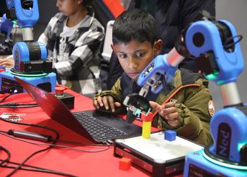 A young dark-haired boy sits at a laptop computer peering at a blue robot arm which is programming through the laptop keyboard