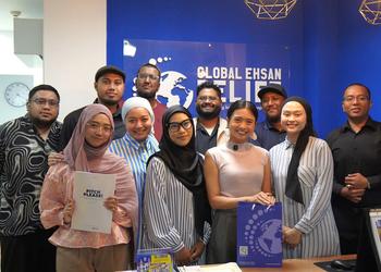 A group of men and women at Muslim anti-poverty charity Global Ehsan Relief stand in a group against a blue wall with their charity logo on it, smiling at the gamera