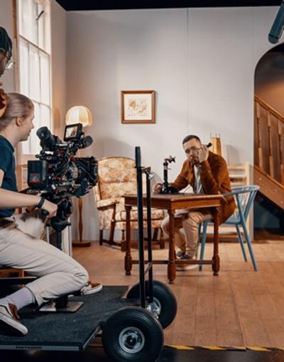 Students using a film camera to film an actor on set who is sitting at a table using a phone
