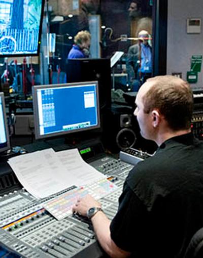 Two individuals in a recording studio, one sitting at a computer and the other standing by a microphone.