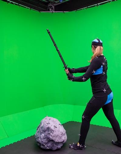 A person in a motion capture suit wielding a prop weapon in front of a green screen