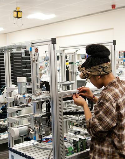 Female student working with a large machine in the MDX mechatronic lab. Two other students are sitting at a table in the background.