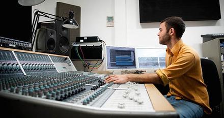 A man sits at a recording studio's mixing desk, adjusting audio levels for a sound production.