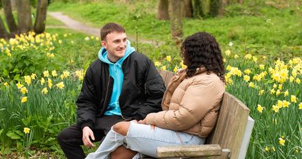 Two students sit on bench with green behind them and daffodils
