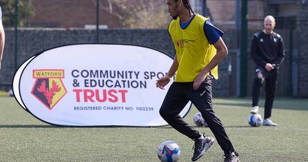 Man controls football on 4G pitch while coach watches on, banner with Watford FC crest reading Community Sport and Education Trust