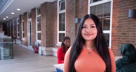 Female student stood on campus with students sat behind