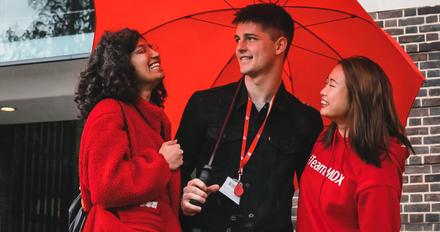 three happy students stand under an umbrella