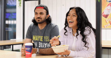 2 middlesex university students, one young man with a beard and one young woman with long brown hair sit at a high table eating lunch