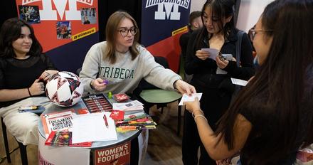 Two students sitting at a stand for Mavericks football, speaking to two other women