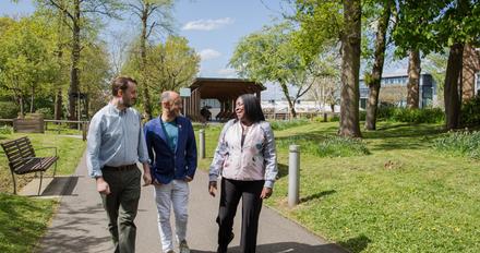 A smiling, diverse group of three staff members are conversing while walking through a green outdoors area on campus.