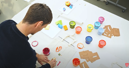 Student working on a white canvas with coloured paint pots on a table