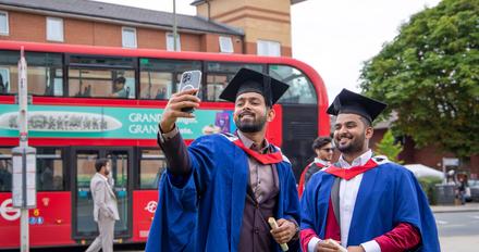 Two smiling male graduates in graduation robes taking a selfie outside College Building