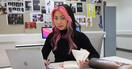 Female student sat at desk looking at book and laptop