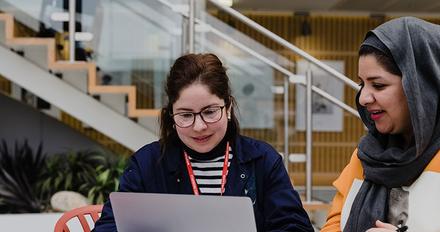 Two women sitting at a table with a laptop, discussing work projects and sharing ideas.