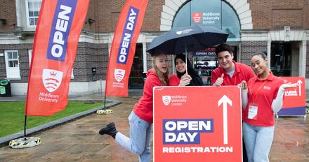 Four smiling students holding an umbrella, behind a sign pointing forwards to open day registration