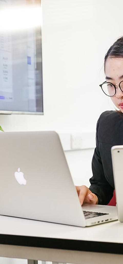 A lady sitting at a desk on her Macbook while holding a bunch of papers in the other hand.