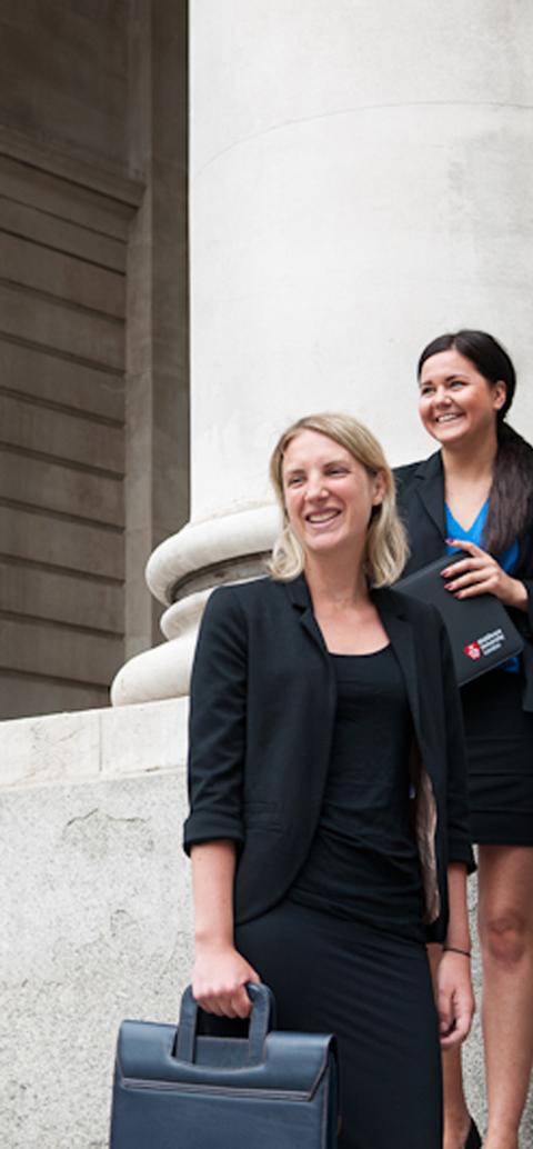 Four professionally dressed students stand outside a building with large stone columns, smiling and holding folders and bags.