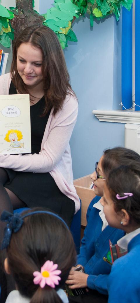 A teacher reads a picture book to a group of primary school children seated in a library.