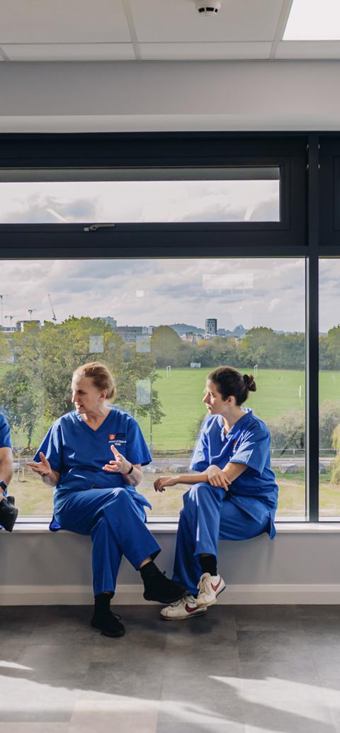 Nursing students in clinical uniforms talk together beside large windows overlooking green space.
