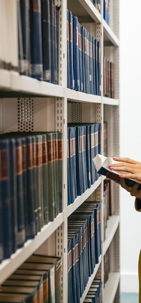 A student reads a book while standing between shelves in a university library.