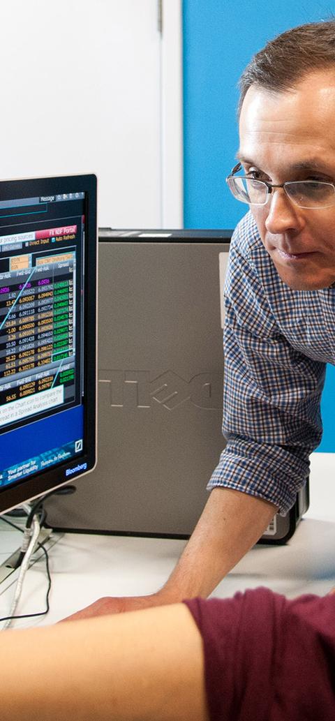A student sits at a desk looking at financial data as a lecturer looks on