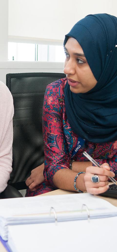 Two female students (one wearing a headscarf) sitting at a desk, talking