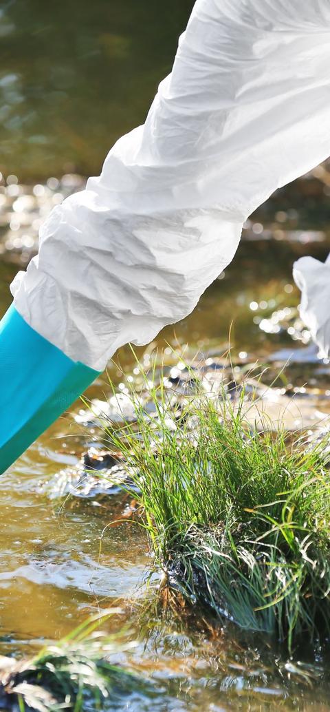 a person in white hazard suit with blue gloves reaches into the water to take a sample