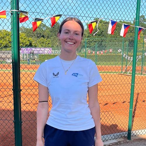 A young woman in a t shirt stands in front of a tennis court