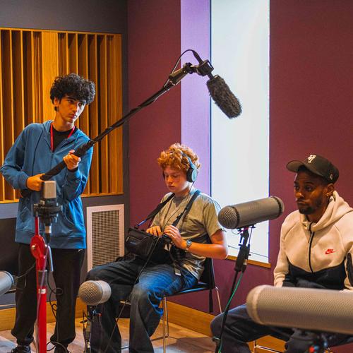 Teenage boy holds a microphone above Middlesex alumnus Jely Kikamba being interviewed for a podcast while another teenage boy listens to the podcast recording