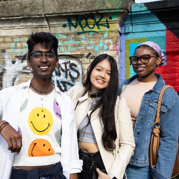Three smiling students in front of a wall with artistic graffiti in Camden