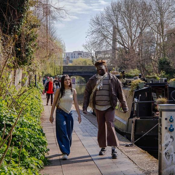 Two MDX students speaking and walking down Regent's Canal near some green leaves and a barge