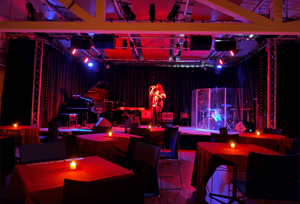 Live band performing on stage in a moody, red-lit music venue with candlelit tables in the foreground