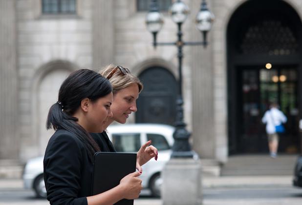 Two students walk and talk outside a university building, carrying study materials.