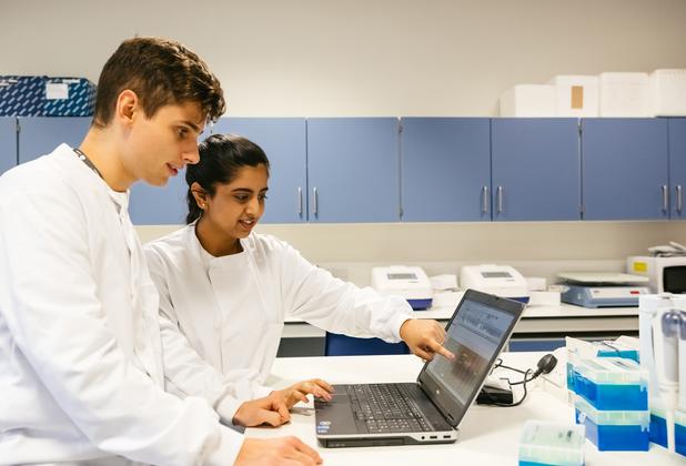 Two students in laboratory coats analysing scientific data together on a laptop in a sciences laboratory.