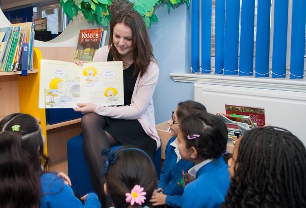 A teacher reads a picture book to a group of primary school children seated in a library.