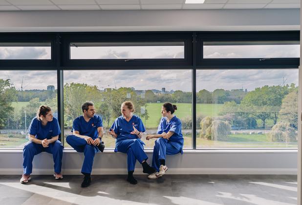 Nursing students in clinical uniforms talk together beside large windows overlooking green space.