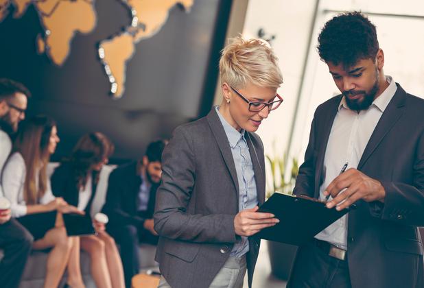 Two colleagues in an office look at a clipboard with colleague sat behind