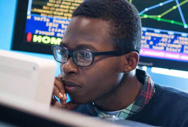 A student sitting looking at a computer screen, with a monitor displaying financial data behind him