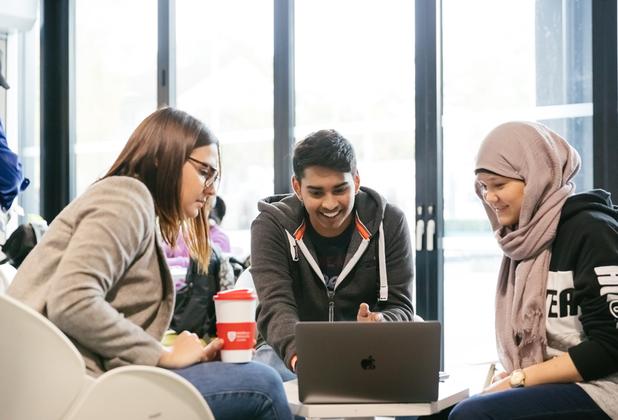 A group of three students gathered round a laptop in a cafe area