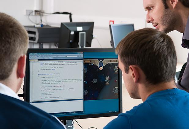 Two men sitting at a desk looking at a computer screen, with a third man standing next to them, leaning over to look at the screen