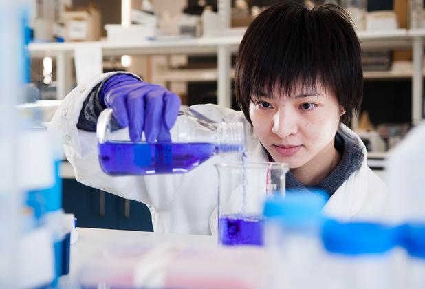 Student in a lab wearing a white coat and blue gloves mixing chemicals