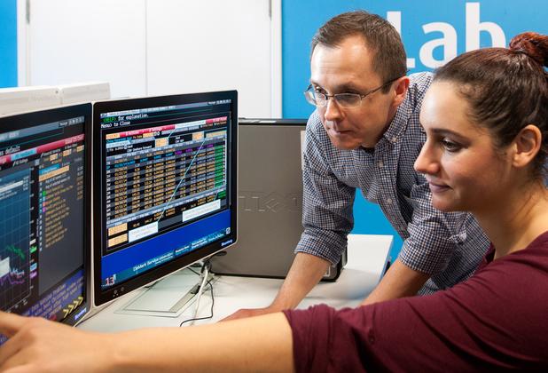 A student sits at a desk looking at financial data as a lecturer looks on